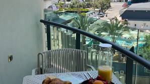 En balkong med ett bord dukat för frukost, med bland annat croissanter, apelsiner och ett glas apelsinjuice, med utsikt över ett semesterområde med en pool och palmer.