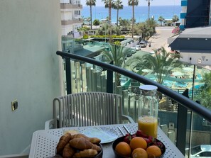 A balcony with a table set for breakfast, including croissants, oranges, and a glass of orange juice, overlooking a resort area with a pool and palm trees. - Qoople Eхсellence B-19 neue Wohnung am Meer by Interhome (Alanya)