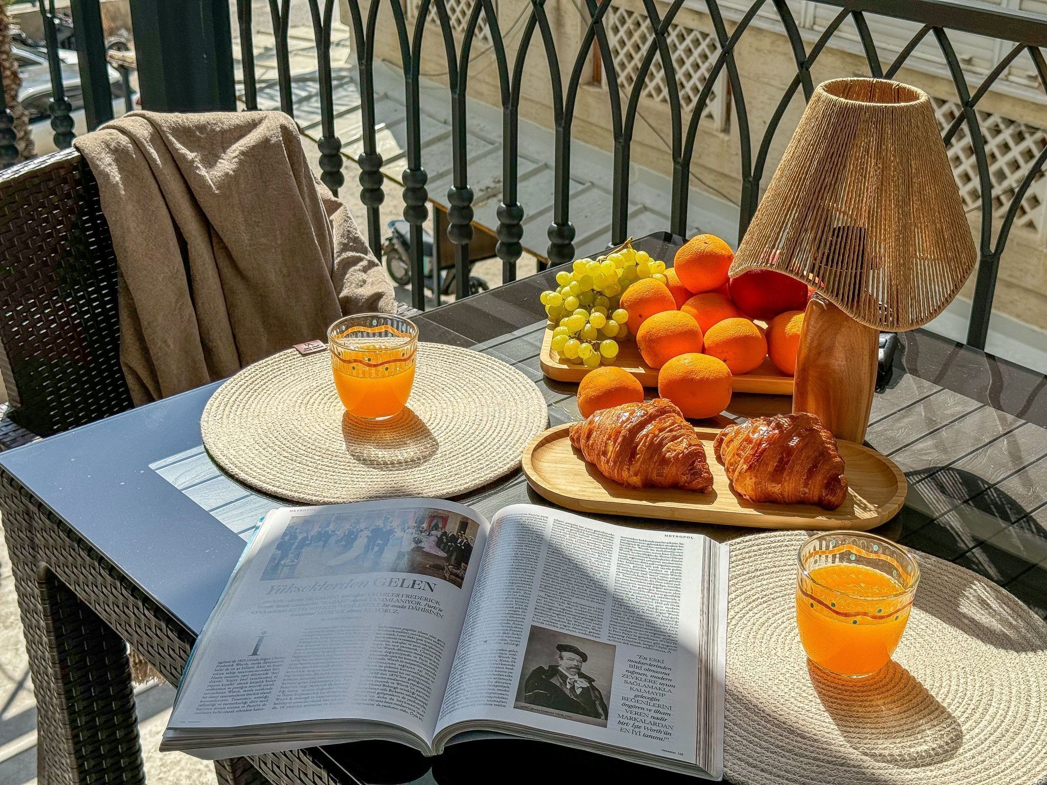 Avoin kirja pöydällä, vieressä lasi appelsiinimehua, lautasella appelsiineja ja rypäleitä, croissant ja kudottu alusta.