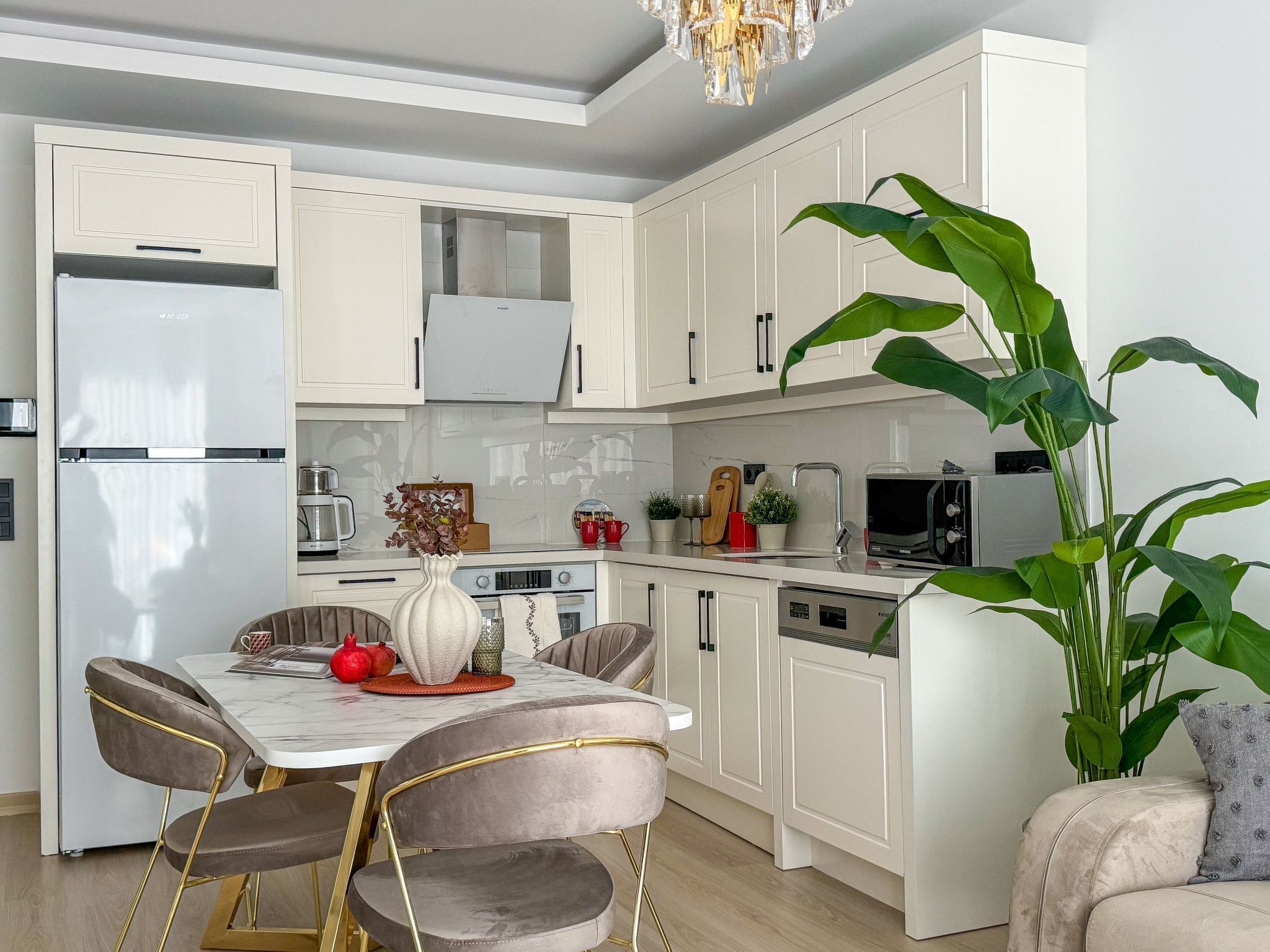Una cocina moderna con armarios blancos, un refrigerador de acero inoxidable, una mesa de comedor con sillas y una planta grande en maceta.