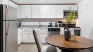 A modern kitchen with white cabinets, stainless steel appliances, a microwave, and a dining area with a wooden table and a vase.
