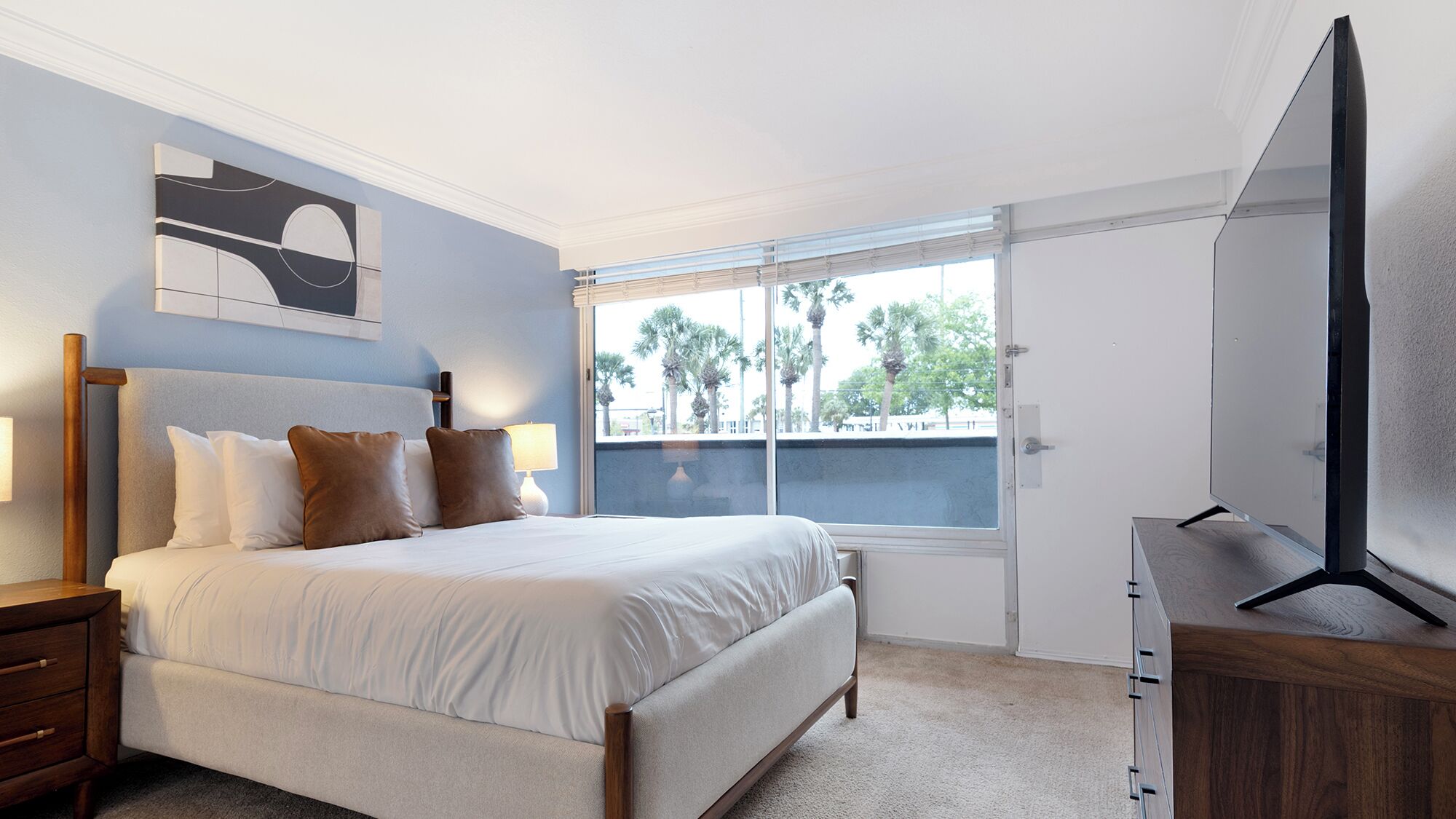 A bedroom with a bed, bedside tables, a TV, a window with a view of palm trees, and a painting on the wall.