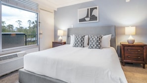 A neatly made bed with a white comforter and patterned pillows, a headboard, two bedside tables with lamps, a window with a view, and a wall-mounted abstract art piece.