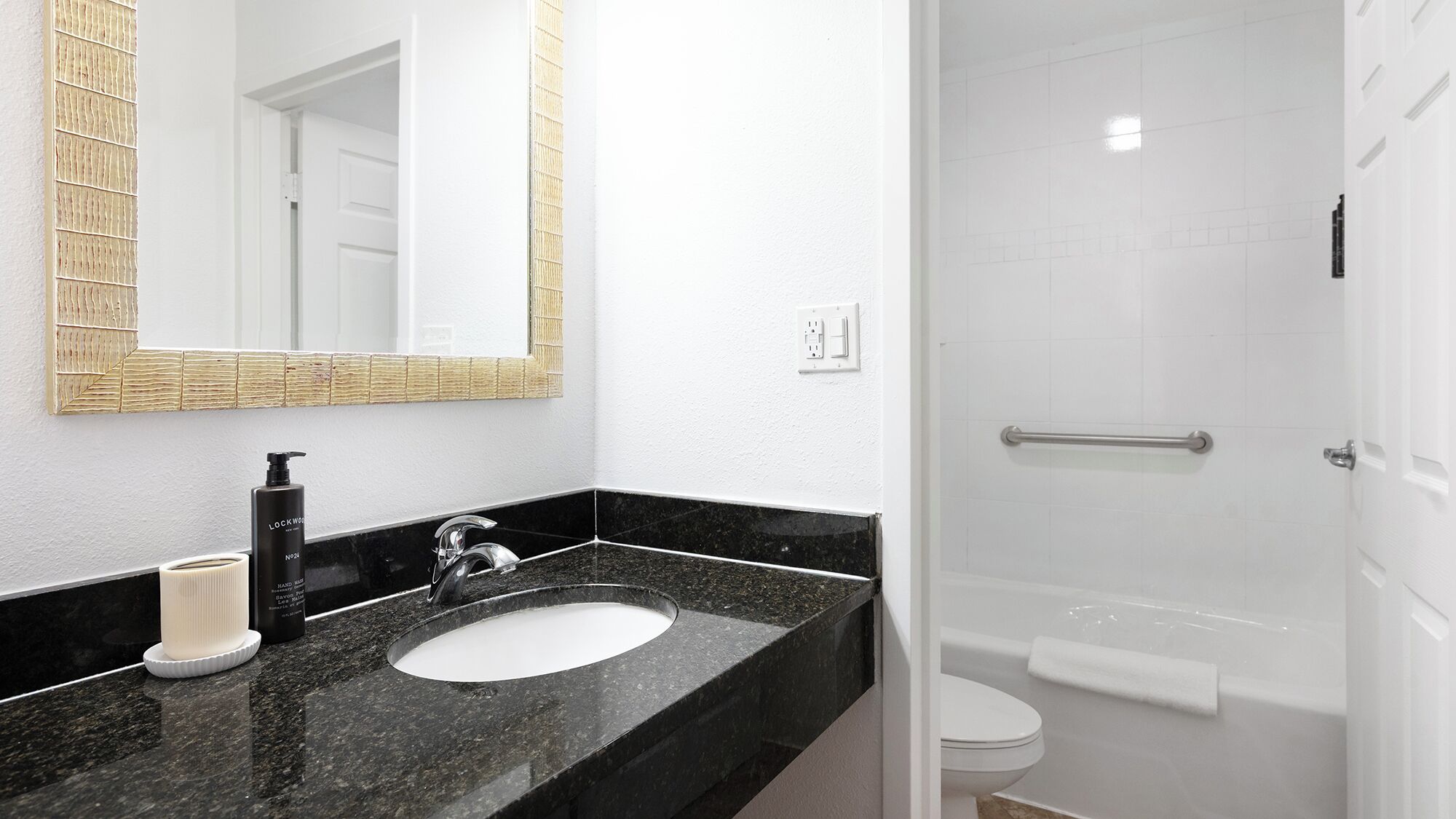 Un bagno con un lavandino in granito nero, uno specchio con cornice dorata, una bottiglia di sapone per le mani e un water bianco.