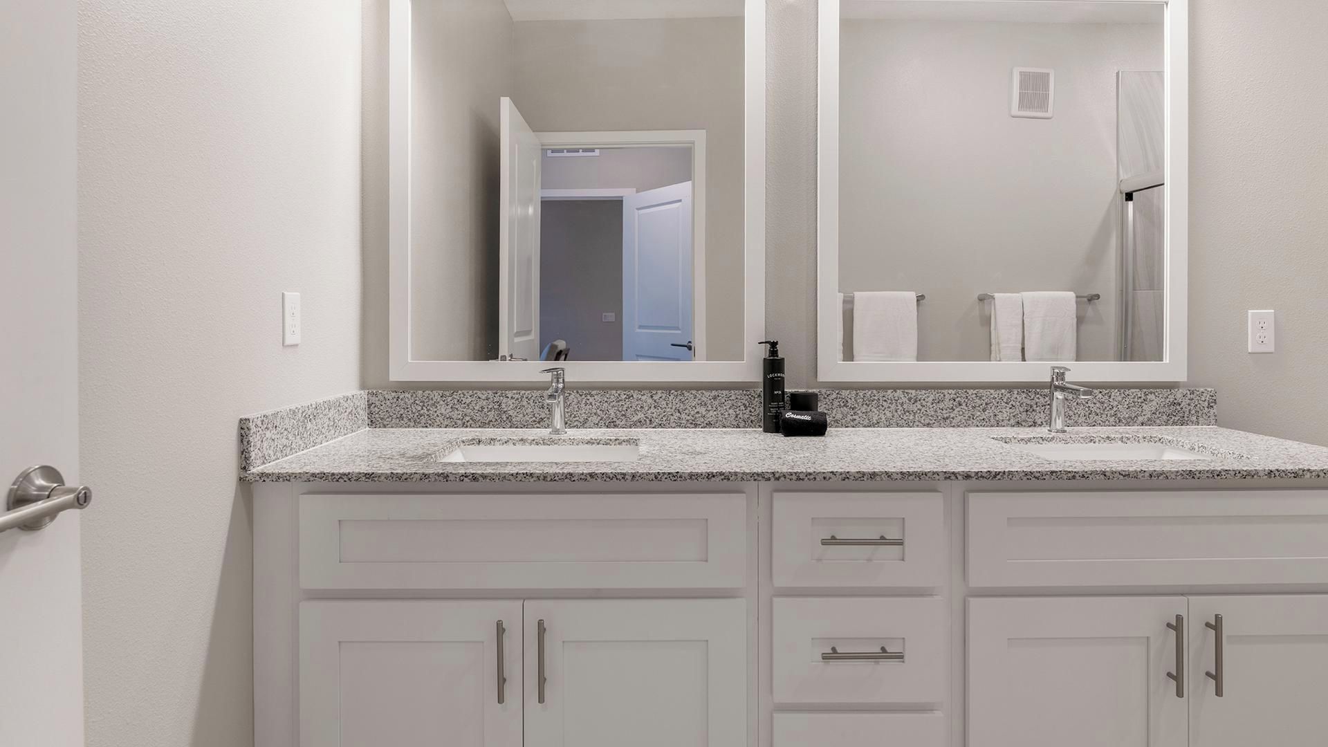 Une salle de bain dotée d’un comptoir en granit, d’un évier double, d’un grand miroir et d’armoires blanches.