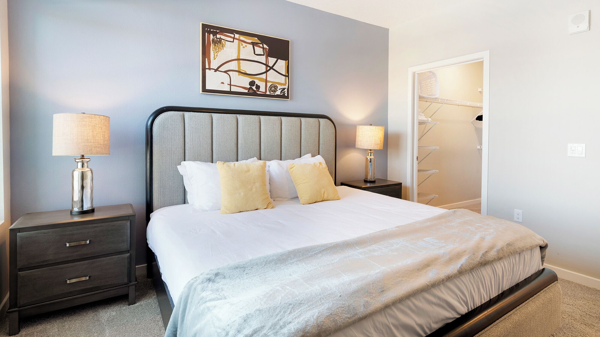 A neatly arranged bedroom with a bed, two bedside tables, a wall-mounted artwork, and a closet with a hanging organizer.