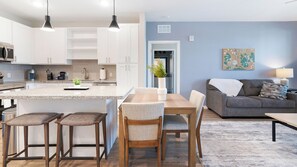 A modern kitchen with white cabinets, a granite countertop, and pendant lighting. There is a dining area with a wooden table and chairs, and a living room with a gray sofa and a rug. - Landing Furnished Apartments Davenport near Northeast Regional Park (Davenport)