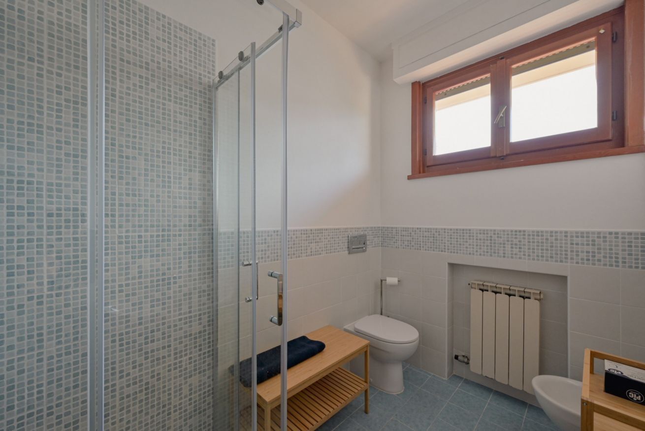 A bathroom with a glass shower enclosure, a wooden bench, a toilet, and a window.