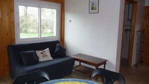 A room with a wooden ceiling, a blue sofa with a cushion, a small table, and a chair. There is a window with a view of trees and a door leading to a shower area. - In a quiet arboretum 3 km from Bergerac town center in Périgord (Bergerac)