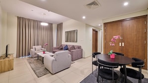 A modern living room with a sofa, armchairs, a coffee table, and a dining table with chairs. There is a TV on a wooden stand, a potted plant, and a large window with curtains. - Grand Stay Holidays Homes Rental Satwa (Dubai)