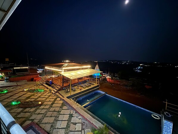 A rooftop pool area with a covered seating area and a view of the city at night. - Exotica De Swiss Panchgini (Mahabaleshwar)