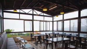 A spacious outdoor seating area with tables and chairs, a sofa, and large windows offering a view of the outdoors. - Exotica De Swiss Panchgini (Mahabaleshwar)