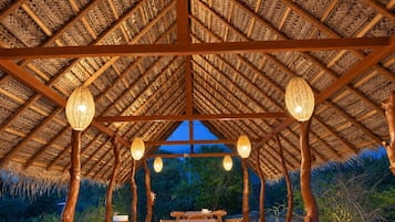 Un espace de restauration en plein air doté d’un toit de chaume, de poutres en bois et de luminaires suspendus. Des tables et des chaises sont disposées pour accueillir les convives.