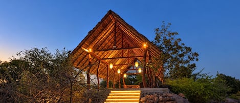 A wooden structure with a thatched roof, surrounded by vegetation and a stone wall.
