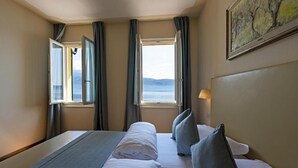 A hotel room with a bed, two windows with curtains, a painting on the wall, and a view of the sea. - Baia d'oro (Gargnano)