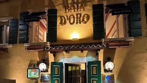 The entrance of a hotel named "Bata D'Oro" with a cobblestone pavement, green shutters, and two potted plants. - Baia d'oro (Gargnano)