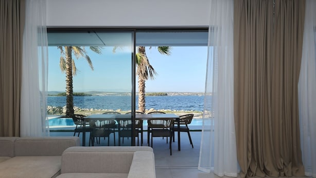 A modern living room with a large glass door leading to a balcony overlooking a body of water and palm trees. - The Palms Resort Villa Waterfront with Pool (Tkon)