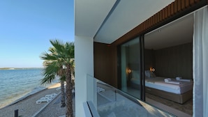 A modern hotel room with a large glass door leading to a balcony overlooking a beach. The room features a bed, a small seating area, and a view of the sea. - The Palms Resort Villa Waterfront with Pool (Tkon)