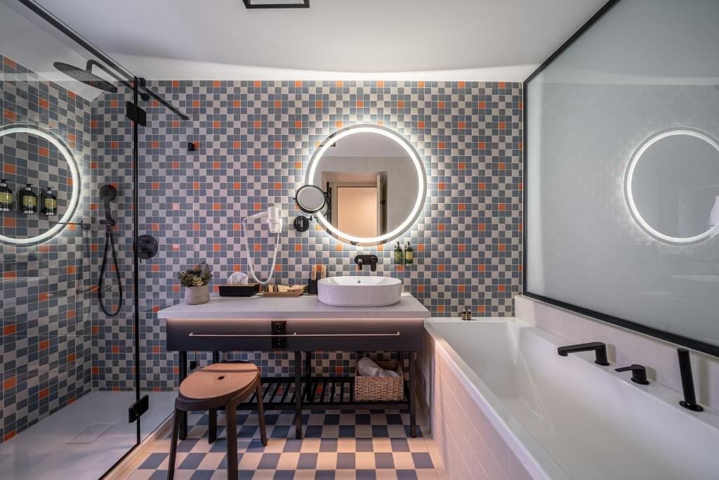 A modern bathroom with a checkered tile backsplash, a round mirror with a light, a white sink, a bathtub, and a shower with a handheld showerhead.