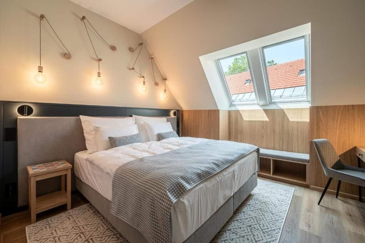 A bedroom with a large bed, a small bedside table, a chair, a window with a view, and a slanted ceiling.