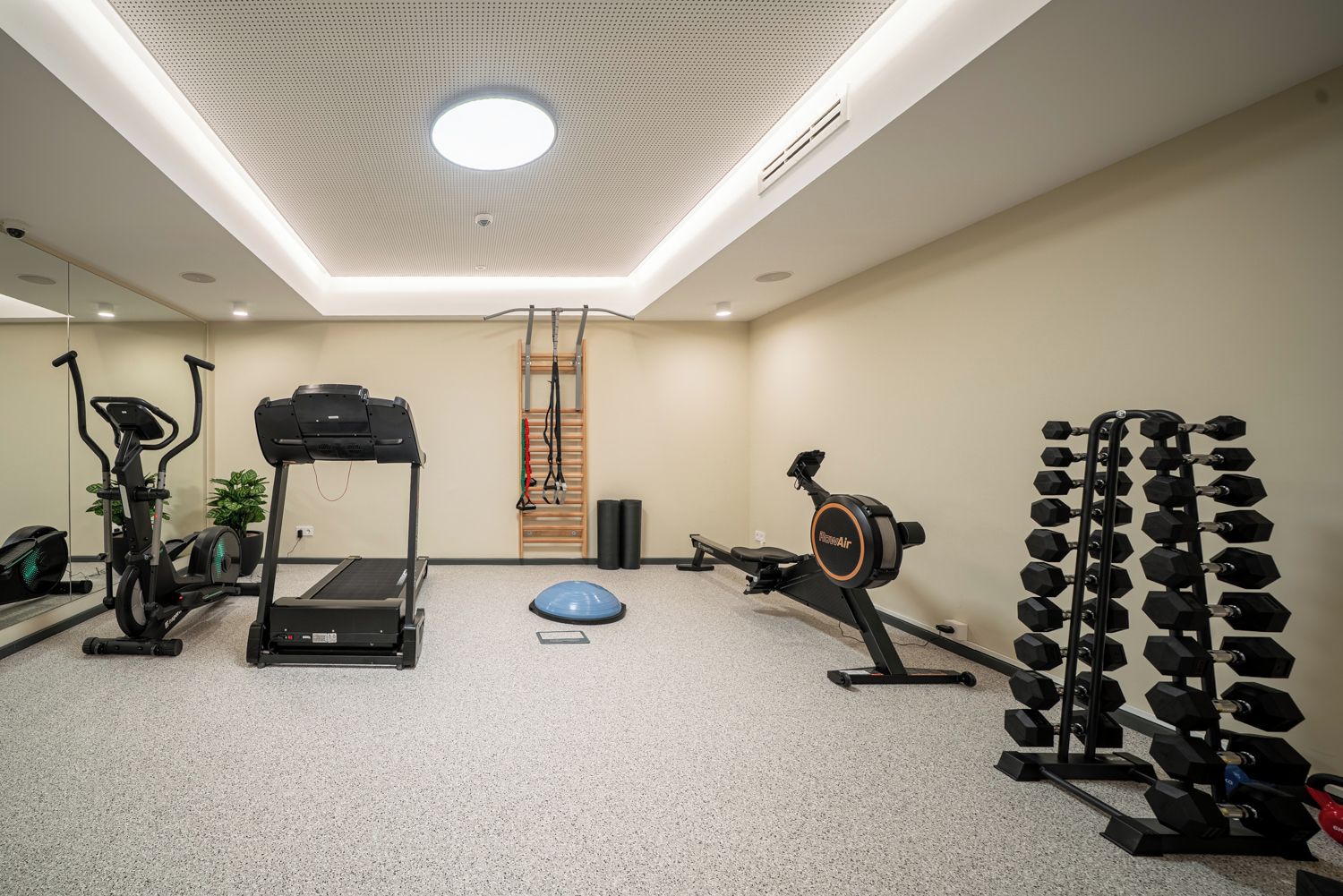 Un gimnasio con equipamiento de ejercicio, incluyendo una cinta de correr, una elíptica, una máquina de remo y un estante de mancuernas.