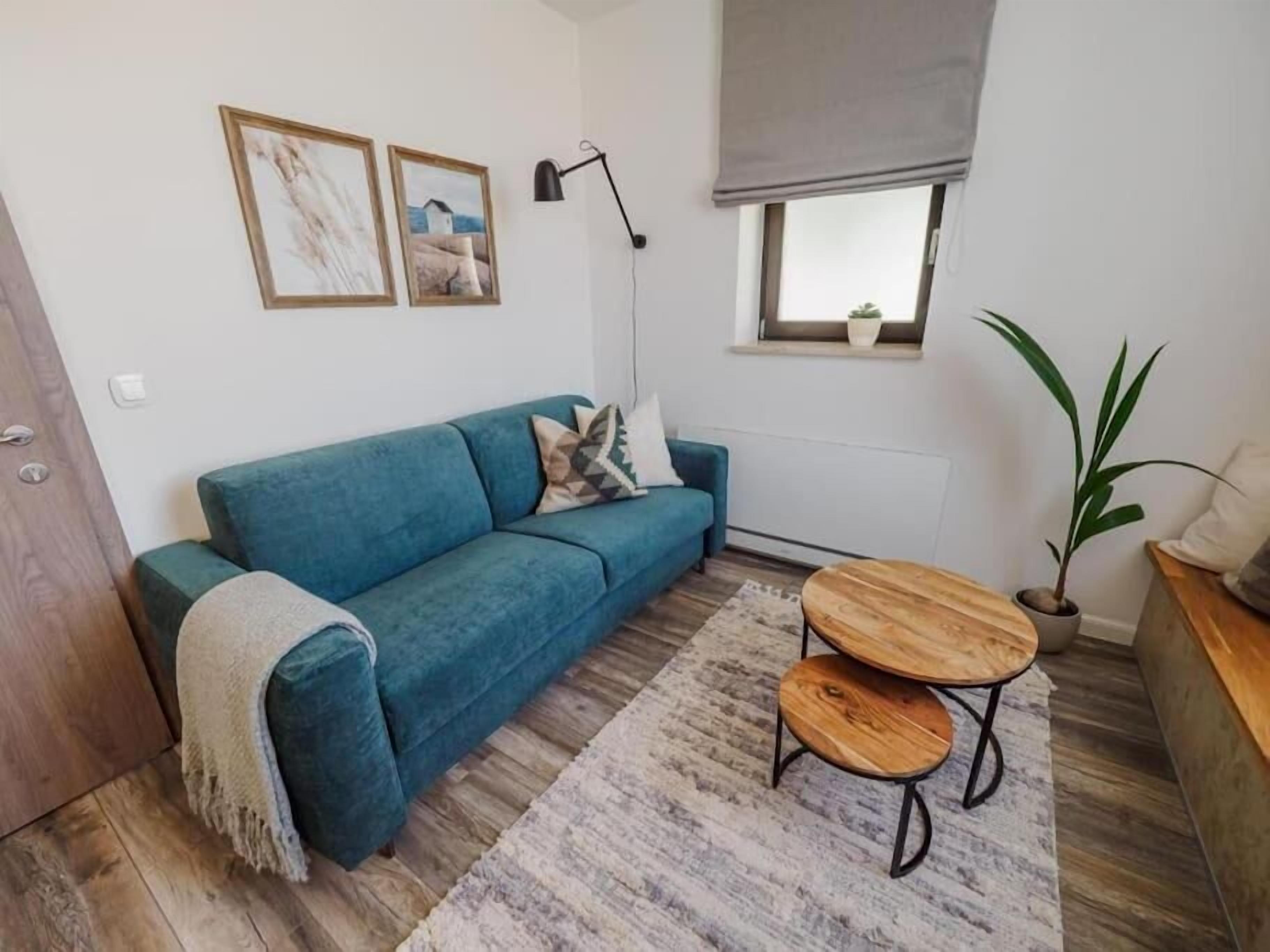 Uma sala de estar moderna com um sofá azul, duas mesinhas de madeira, um abajur e um vaso de plantas.