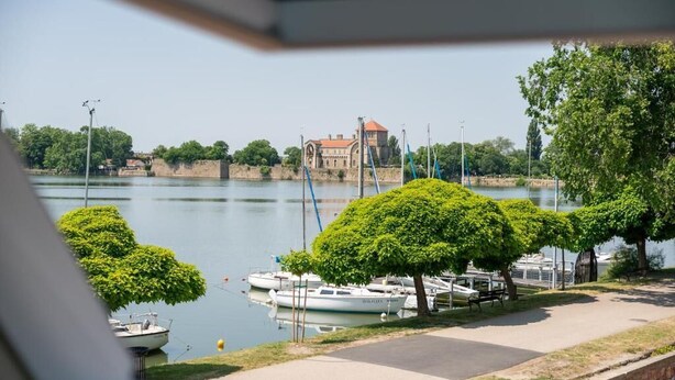 A view of a calm river with boats docked, a historic building in the background, and well-maintained greenery along the riverbank. - Malom és Kacsa Boutique Hotel (Tata)