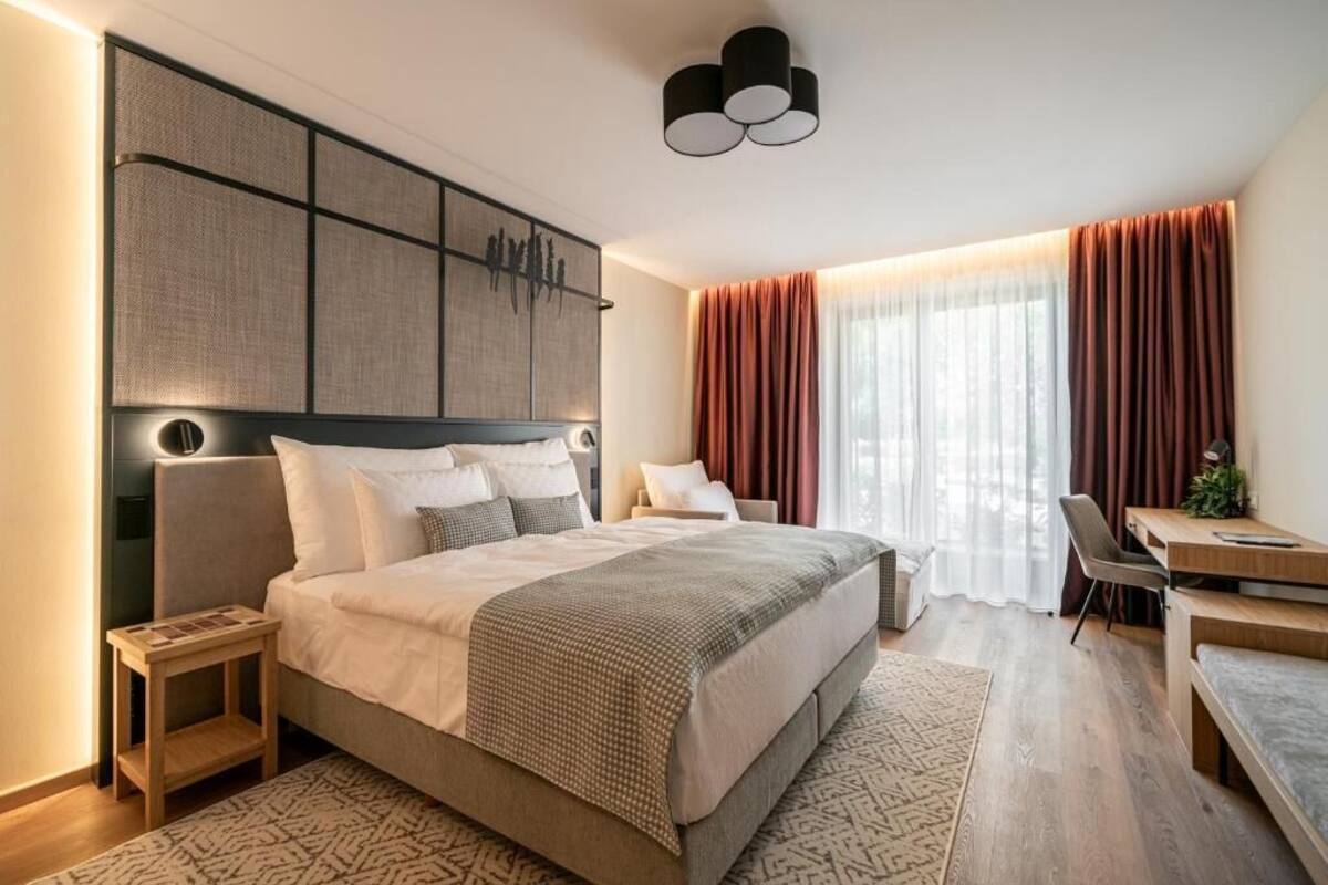 A modern hotel room with a large bed, a desk, and a chair. There is a large window with curtains, a wall-mounted artwork, and a ceiling light fixture.