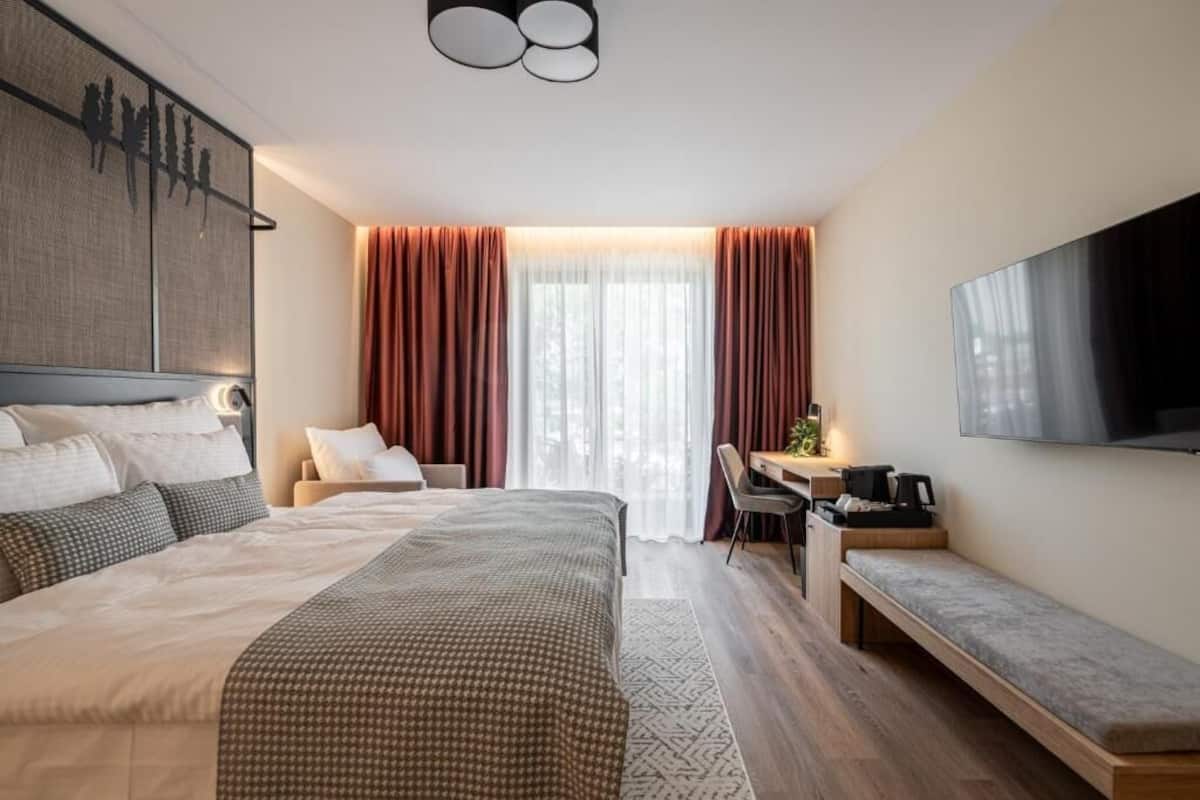 A modern hotel room with a large bed, a desk with a chair, a bench, and a flat-screen TV mounted on the wall.
