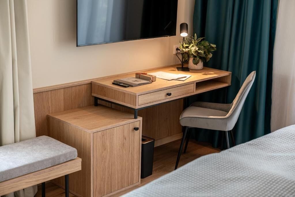 A compact hotel room with a wooden desk, a chair, a bedside table, a small trash bin, and a bed.