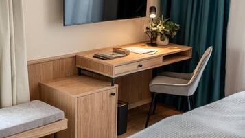 A compact hotel room with a wooden desk, a chair, a bedside table, a small trash bin, and a bed.