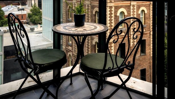 Un balcon avec deux chaises et une table, d’où l’on peut admirer un bâtiment historique doté d’arcatures et d’une tour horlogée.