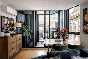 A modern interior with a dining area, a sofa, and a view of the cityscape through large windows. - StayCentral - The Harmony @ Collingwood - Robert St, Collingwood - 2 bedrooms, 2 Beds, 1 Bathroom, 1 Parking (Collingwood)