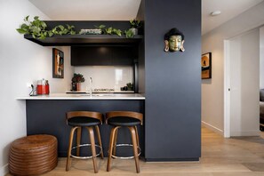 A modern kitchen with a dark wall, two wooden bar stools, a small round wooden stool, and a Buddha statue. - StayCentral - The Harmony @ Collingwood - Robert St, Collingwood - 2 bedrooms, 2 Beds, 1 Bathroom, 1 Parking (Collingwood)