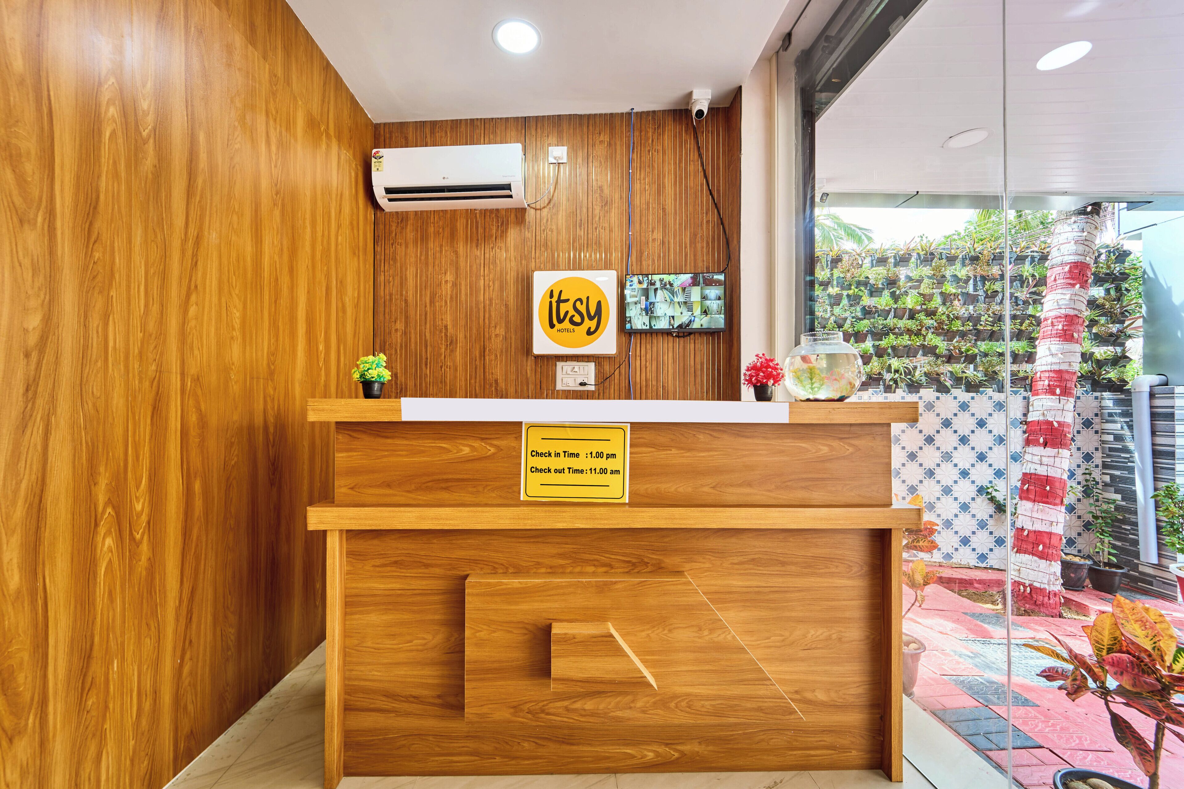 A wooden reception desk with a sign indicating check-in and check-out times, a mounted television, and a wall of potted plants.