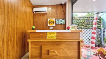 Un mostrador de recepción de madera con un cartel que indica horarios de registro de entrada y salida, un televisor montado y una pared de plantas en macetas.
