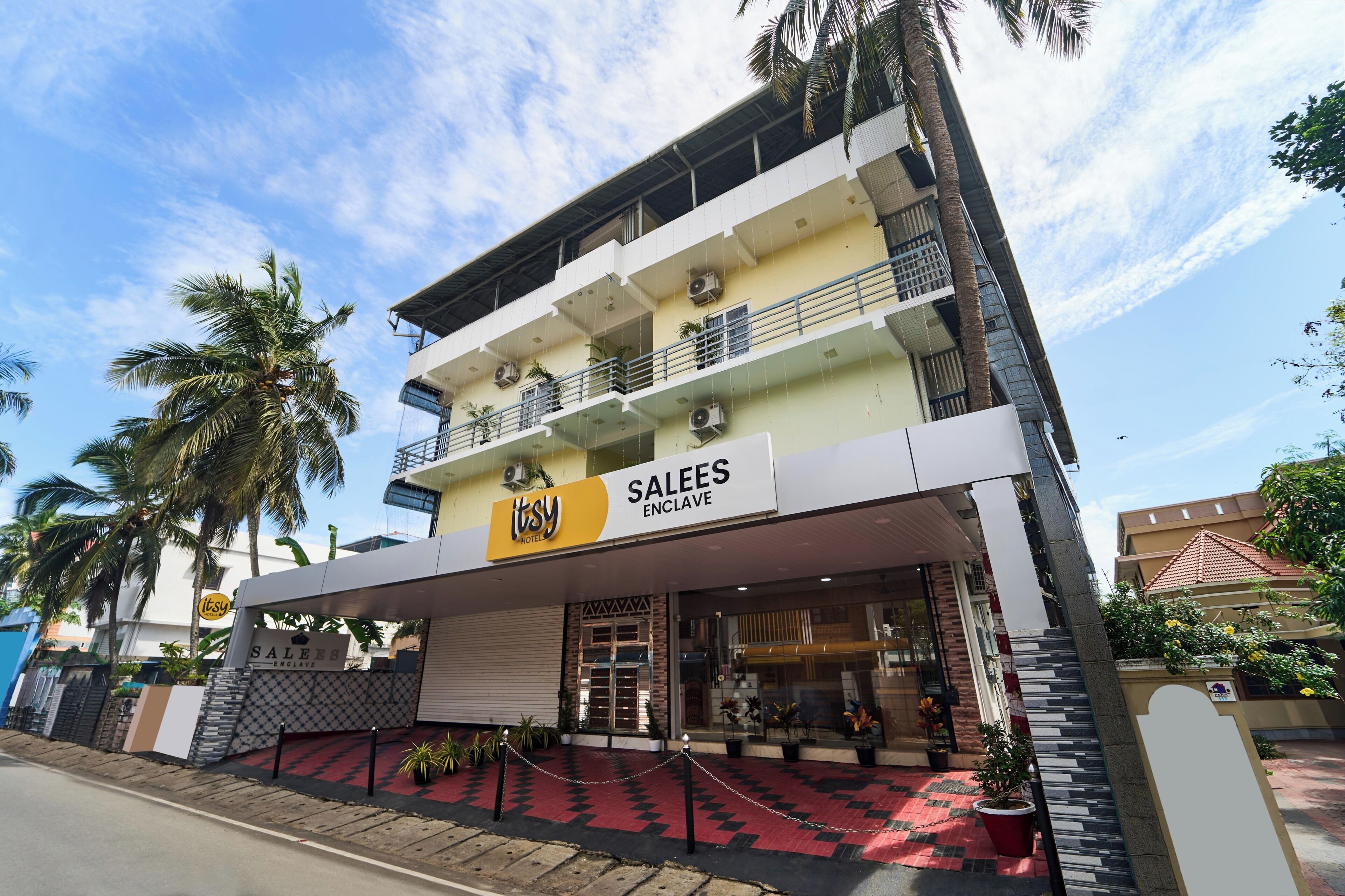Un hotel llamado 'Itsy' con un letrero 'SALEES ENCLAVE', que presenta una estructura de varios pisos con balcones, una entrada protegida por una reja y un suelo con diseño rojo y negro.