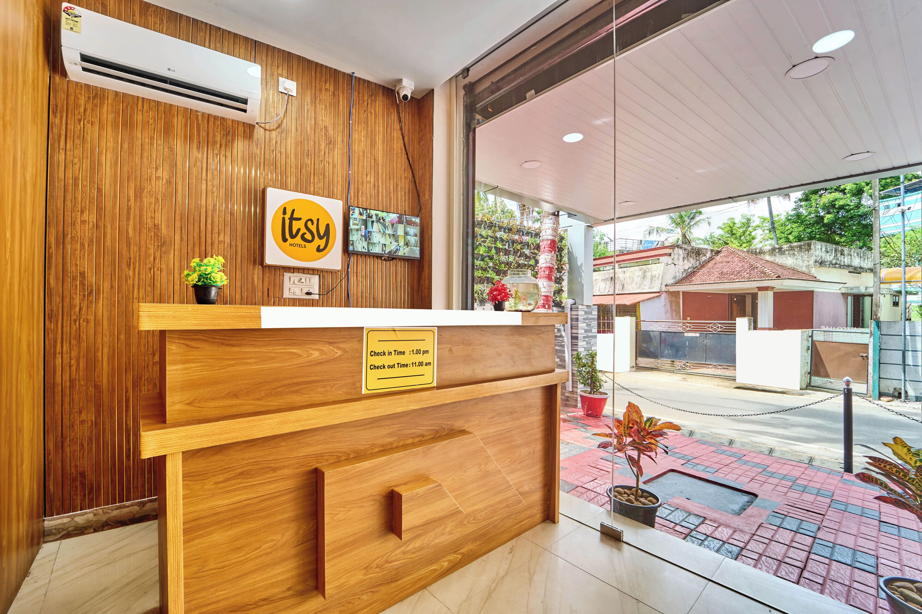 A hotel reception desk with a wooden finish, a sign indicating check-in/out times, and a view of the street outside.