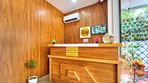 Un mostrador de recepción de madera con un letrero de registro de entrada/salida, un equipo de aire acondicionado, un televisor y vistas a un patio empedrado con plantas.