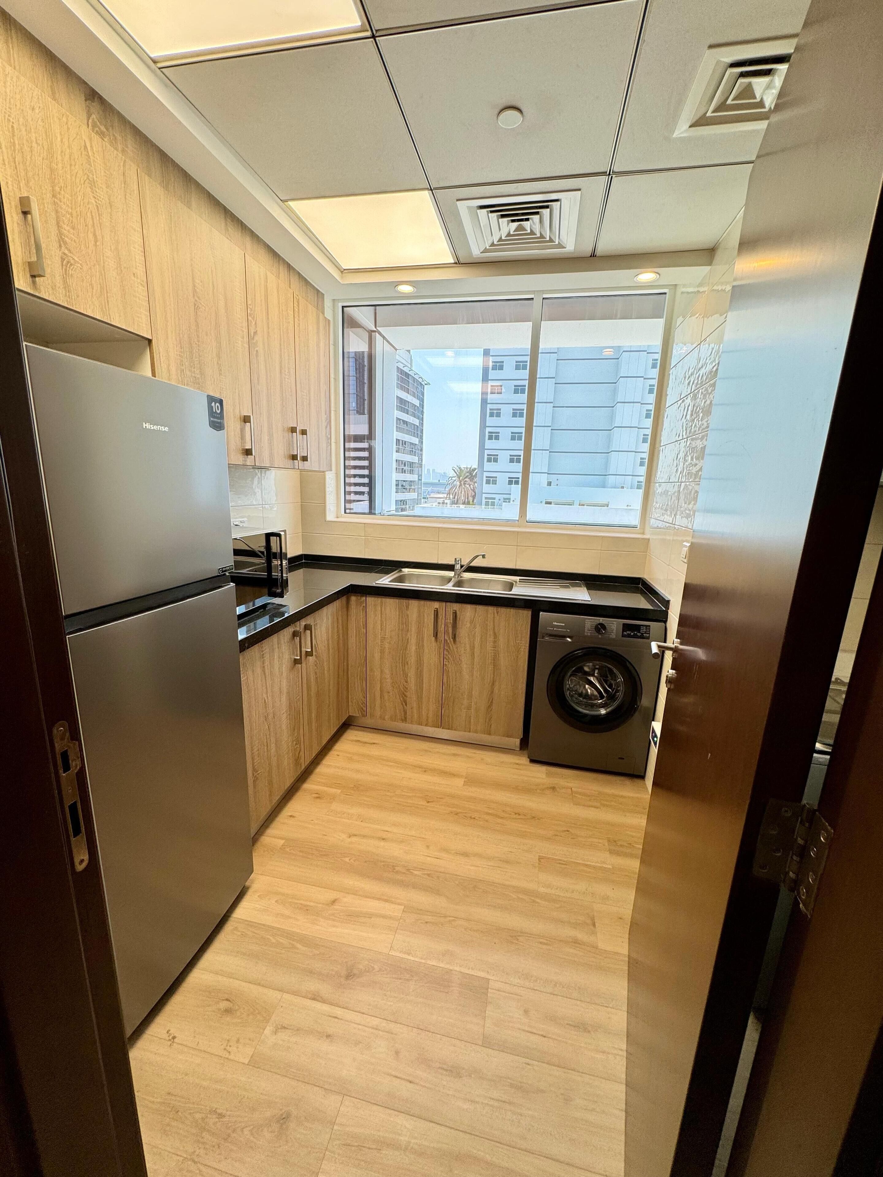 A compact kitchen with a stainless steel refrigerator, wooden cabinets, a washing machine, and a window with a city view.