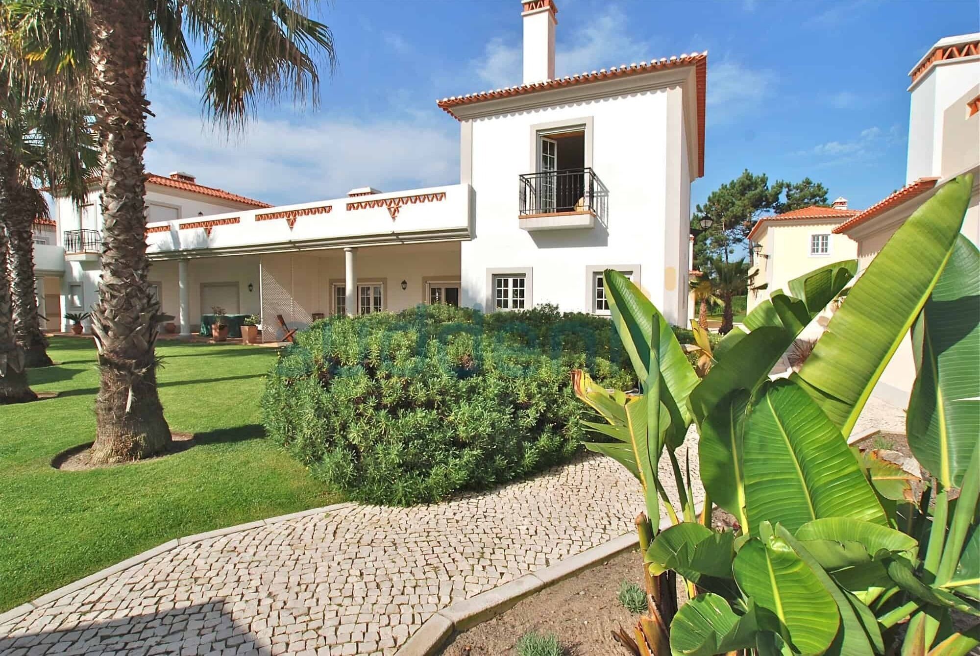 A white building with a tiled roof, a balcony, and a paved pathway leading to a green lawn.