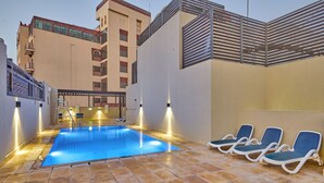 Ein Schwimmbecken mit Liegestühlen und einer überdachten Sitzgruppe, umgeben von einem Mehrstöckiger Gebäude mit Balkonen.