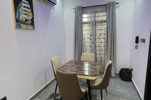Un coin repas avec une table rectangulaire et quatre chaises, un tableau au mur, une fenêtre avec des rideaux et un téléphone fixé au mur.