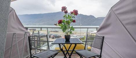 A balcony with a table and chairs, a potted plant, and a view of a body of water and mountains.