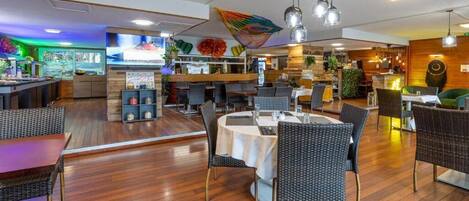 Un restaurante con mesas redondas y sillas de mimbre, una zona de bar y una pantalla de televisión mostrando comida.