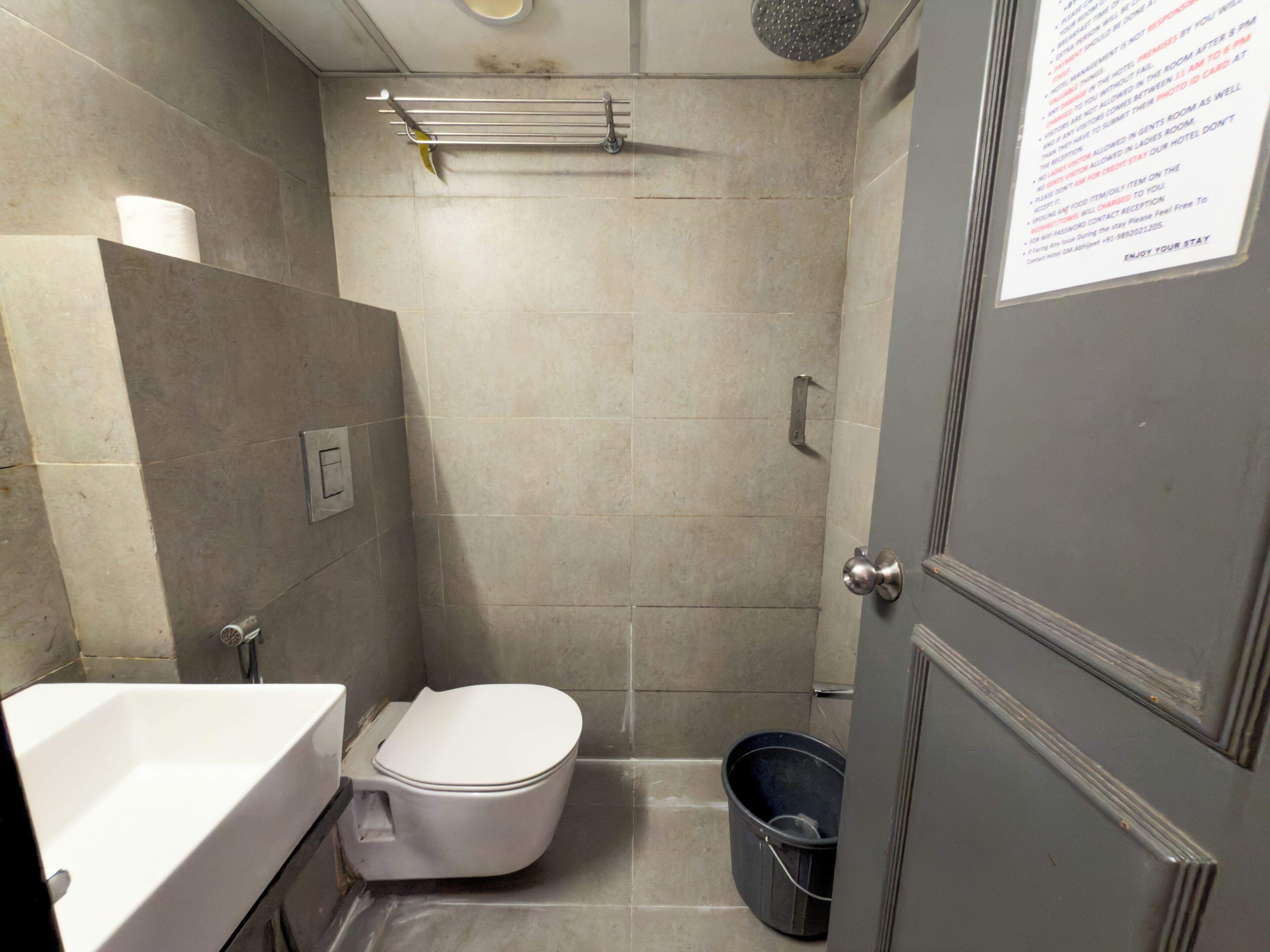 Une petite salle de bain carrelée, équipée de toilettes, d’un lavabo et d’une douche. On aperçoit un porte-serviettes au-dessus des toilettes et un seau posé sur le sol. Une porte avec une pancarte est visible sur la droite.