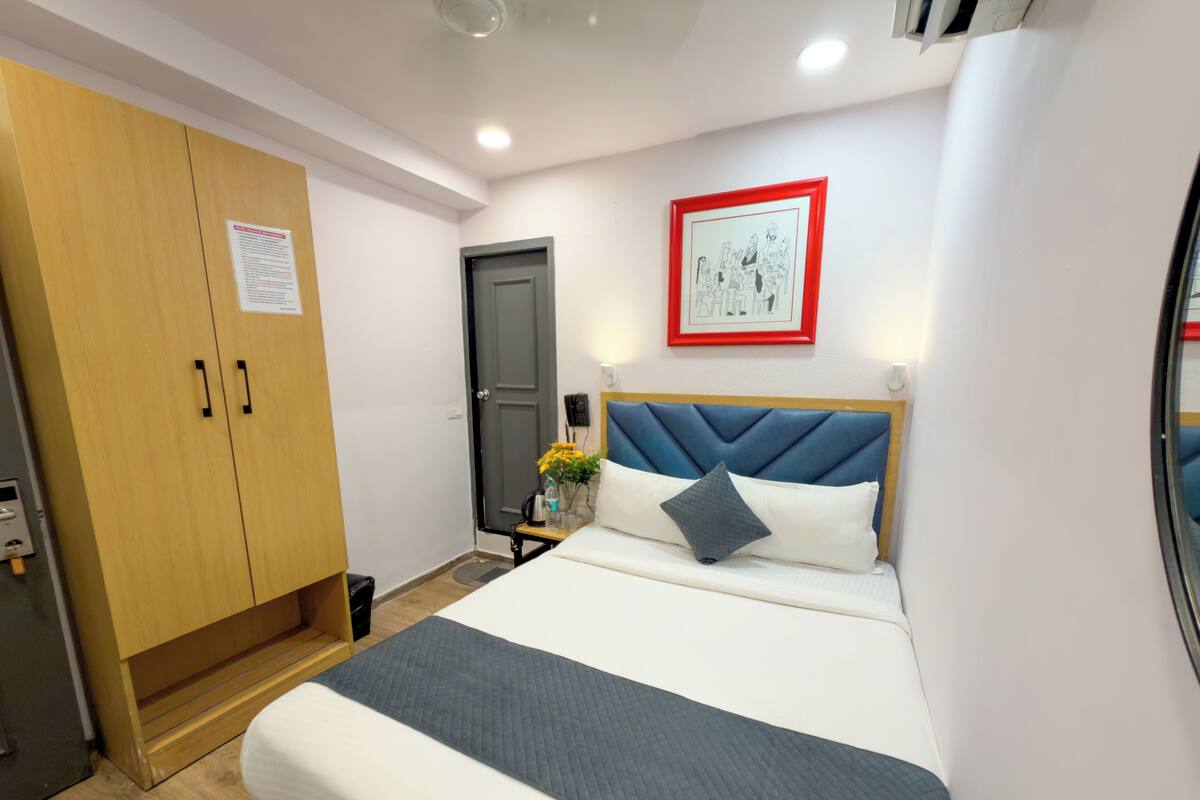 Ein Hotelzimmer mit einem Bett, einem Kleiderschrank, einem Nachttisch mit Blumenvase, einem gerahmten Bild an der Wand und einer Tür.