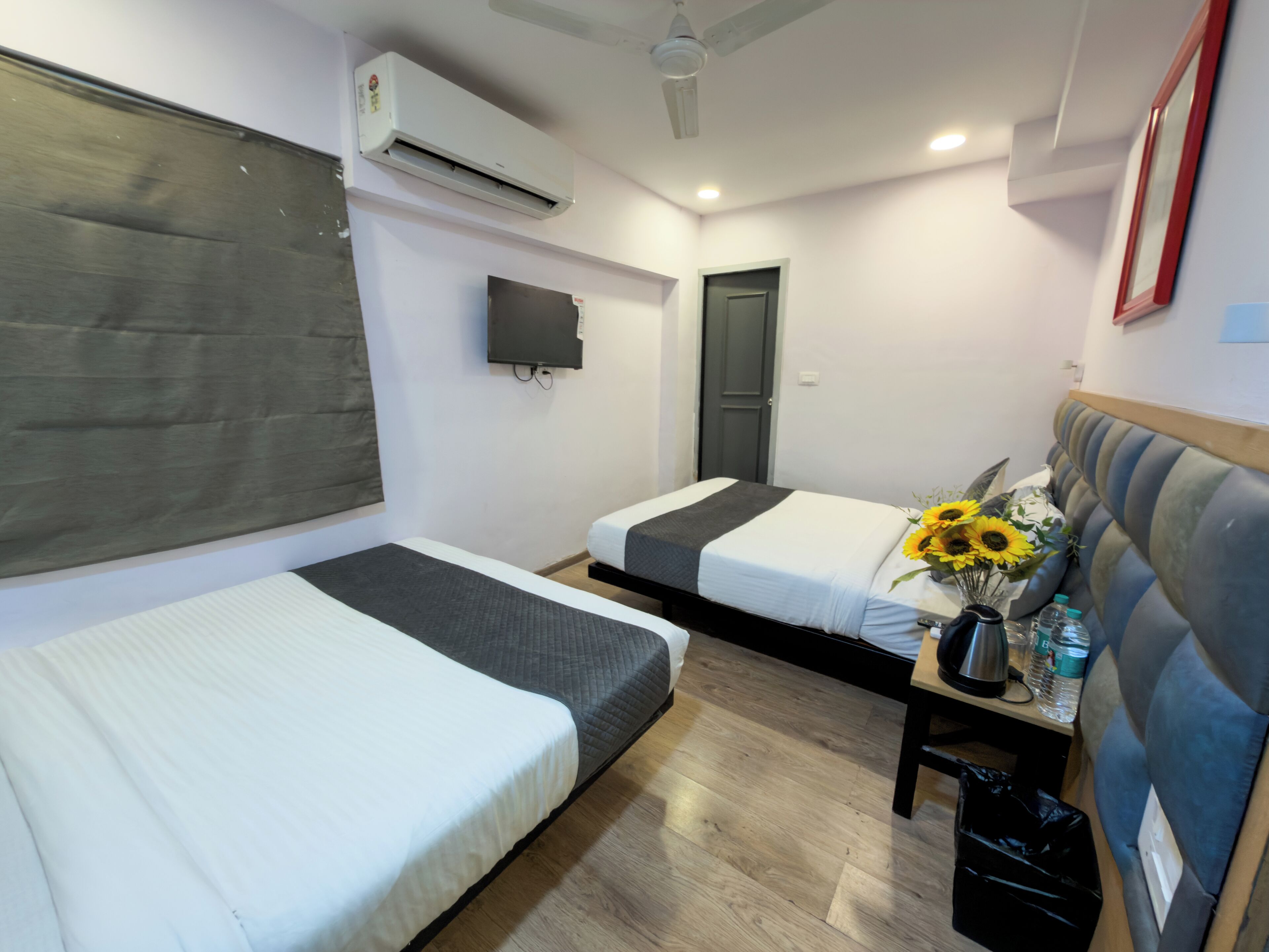 Habitación de hotel con dos camas, televisión, aire acondicionado, piso de madera y una pequeña mesa con un jarrón de girasoles.
