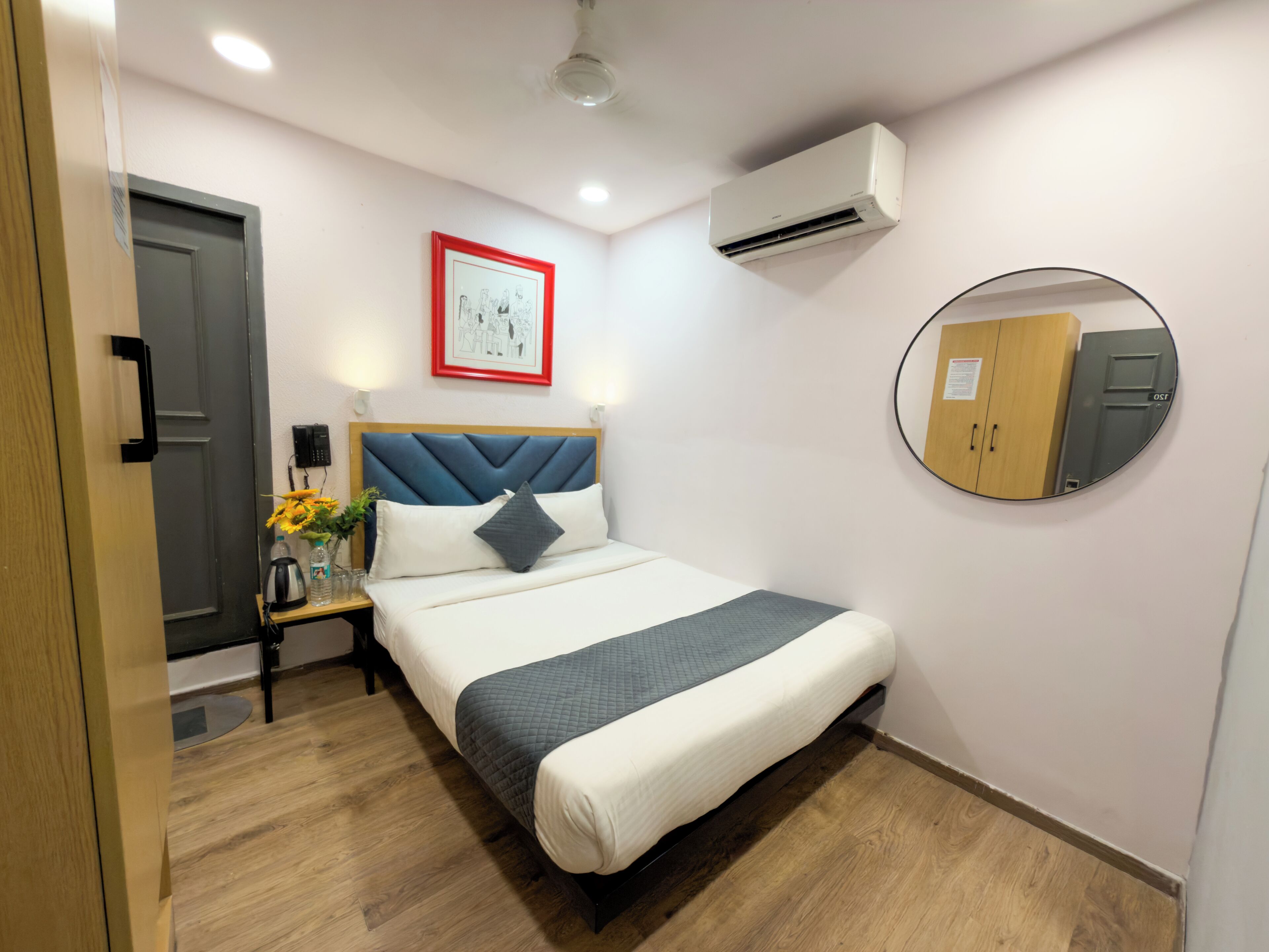 A hotel room with a bed, a bedside table with a vase of flowers, a wall-mounted air conditioner, a framed picture, a round mirror, and a wardrobe.