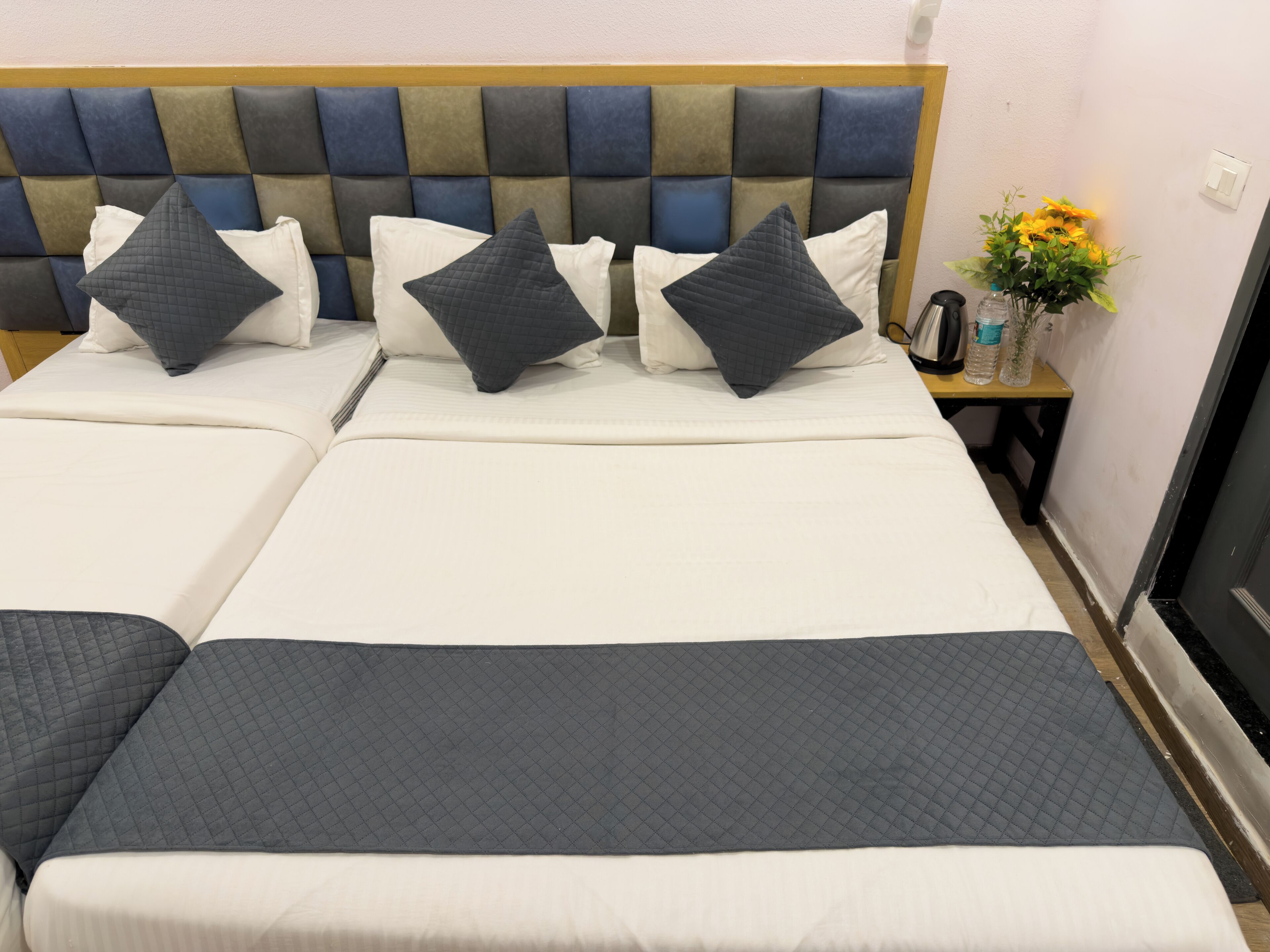 A double bed with a headboard, two pillows, a bedside table with a vase of flowers, and a wall socket.